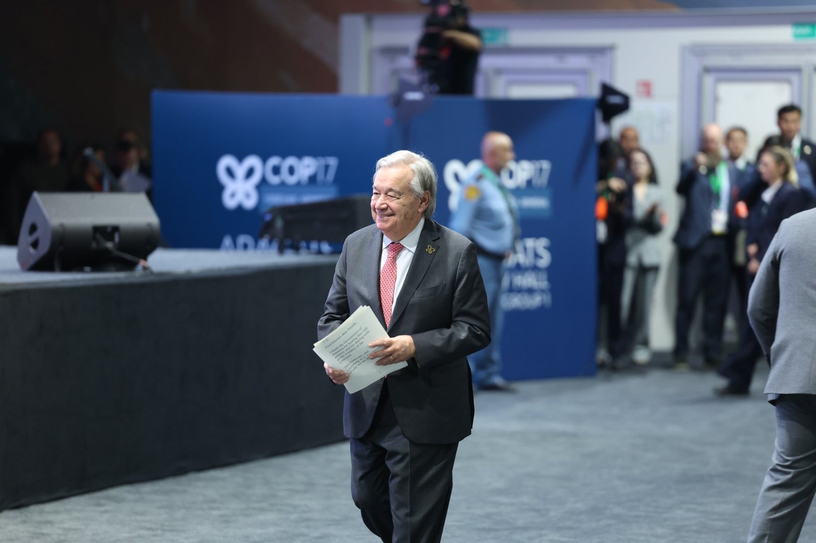 UN Secretary-General António Guterres smiling and walking across the hall, holding the pages of his speech, in front of COP17 banners