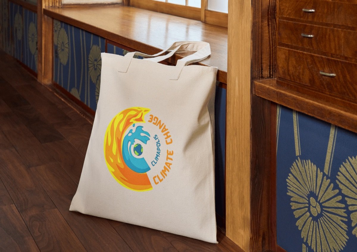 A Climapolis canvas tote bag resting on a wooden bench, printed with the project emblem: a swirl of fire and water circling a globe, ringed by the words Climate Change