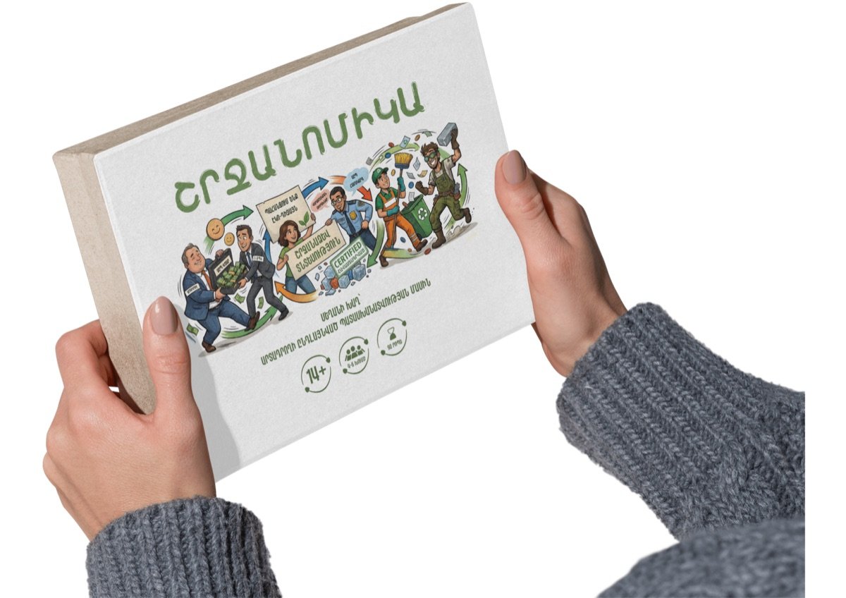 Two hands holding the Shrjanomics game box, titled ՇՐՋԱՆՈՄԻԿԱ, illustrated with producers, officials and waste workers, and marked 14+